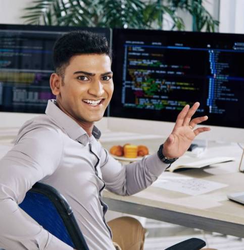 Happy excited Indian software developer sitting at office desk, turning back and waving at camera
